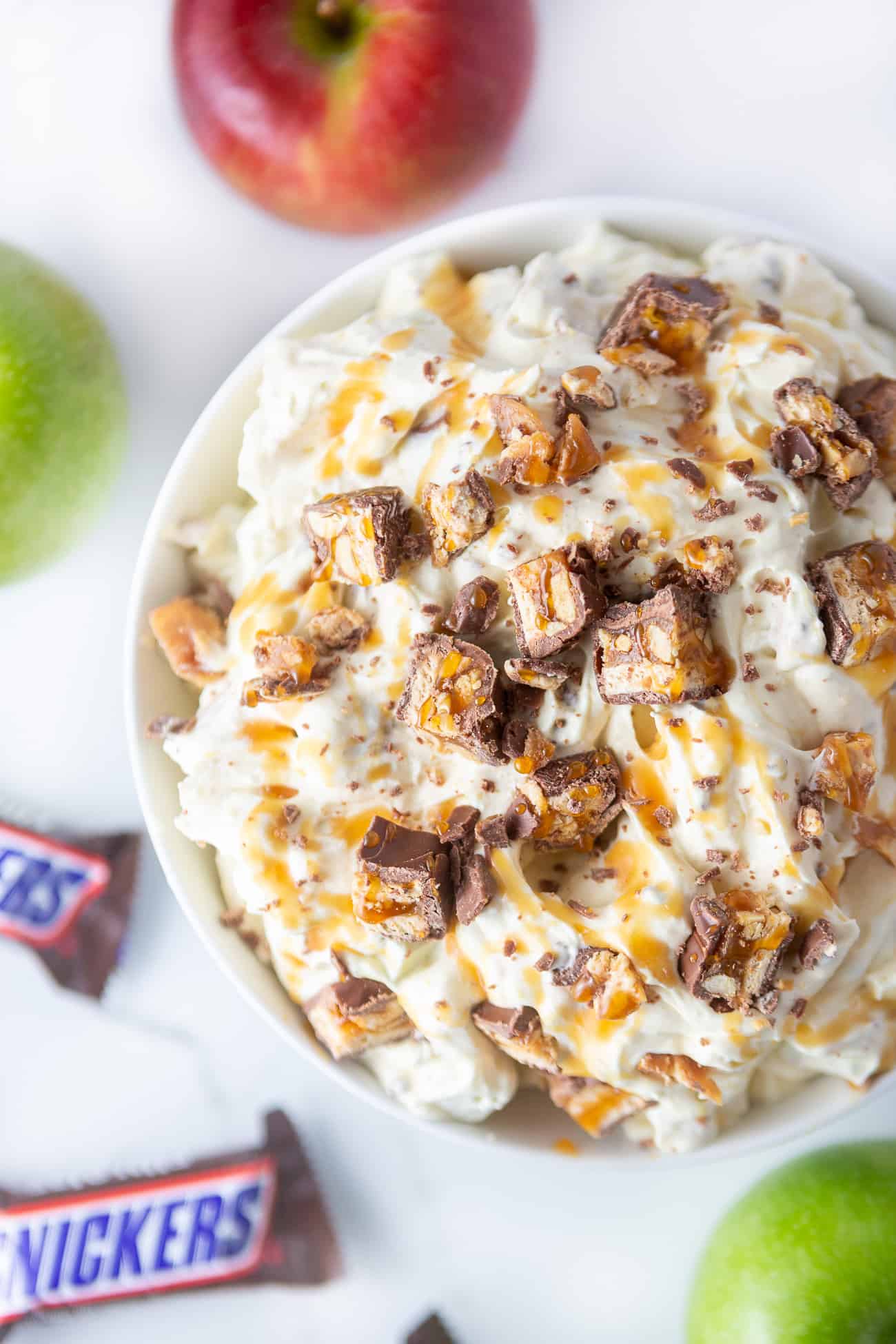 White bowl of apple snickers salad. A fluffy vanilla pudding dessert topped with chopped snickers candy bars and caramel sauce.