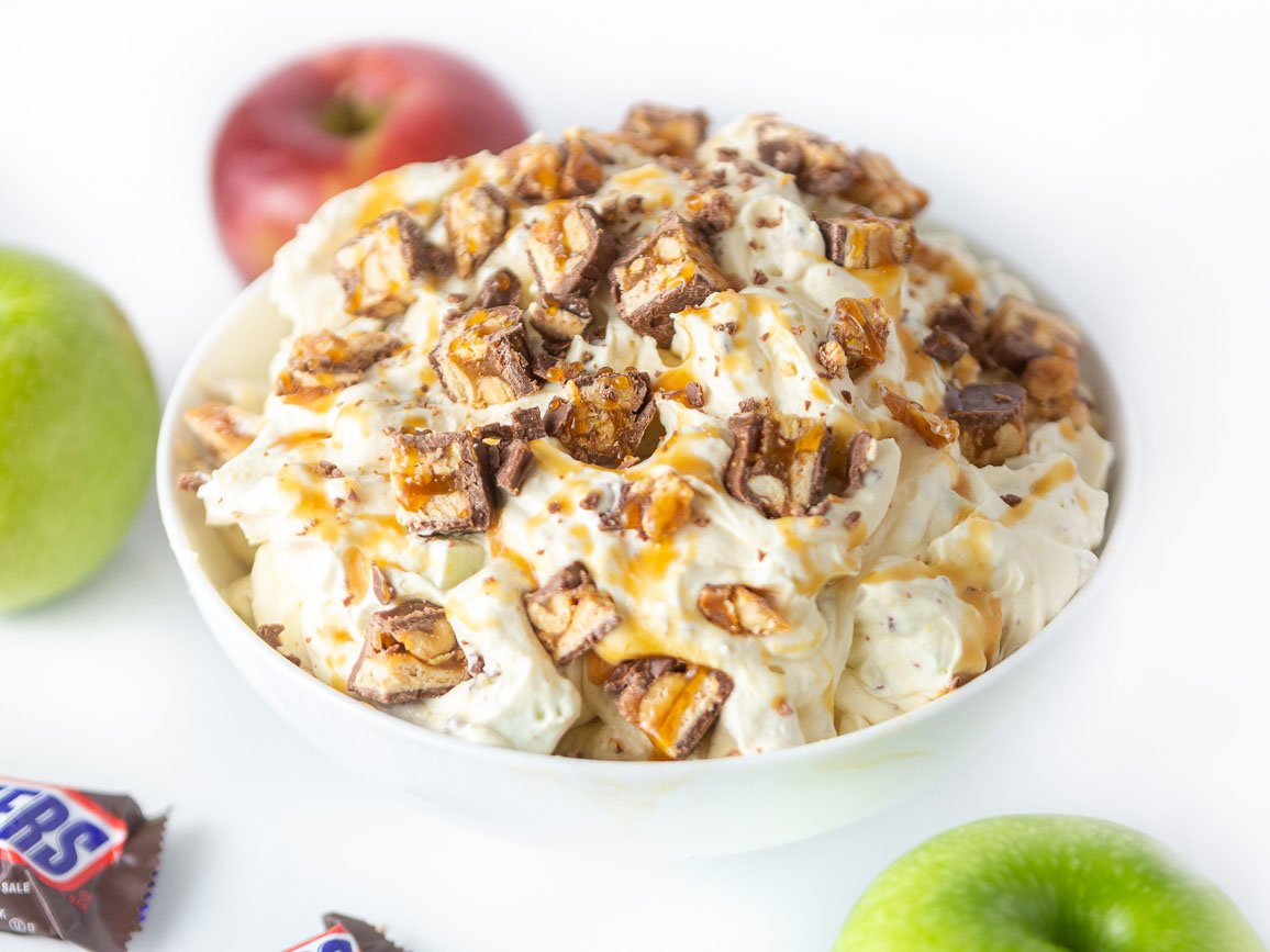 Bowl of creamy apple snickers salad dessert, topped with chopped snickers candy bars, apples, and caramel sauce