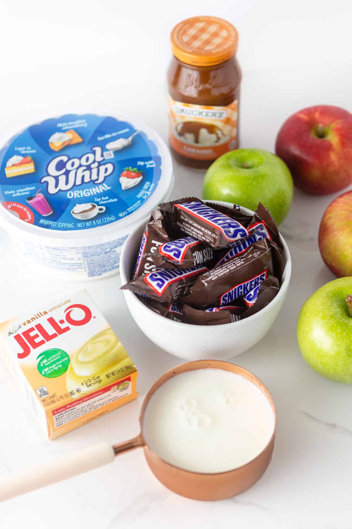 Ingredients for apple snickers salad; includes bowl of snickers, cool whip, a box of instant vanilla pudding mix, milk, apples, and a jar of Smuckers caramel sauce