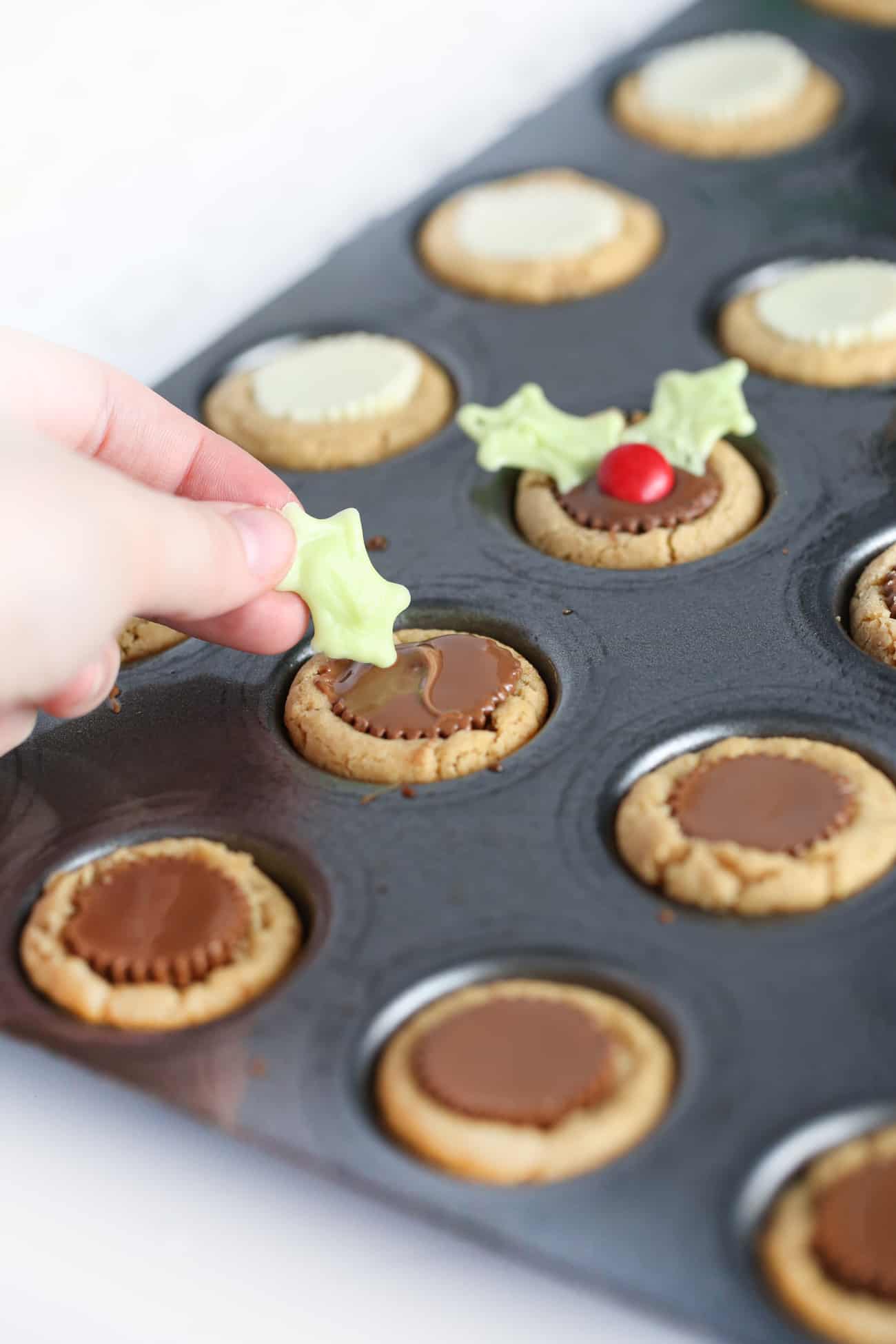 Hand placing white chocolate holly leaf into Reeses peanut butter cup cookie to make easy Christmas holly cookie cups.
