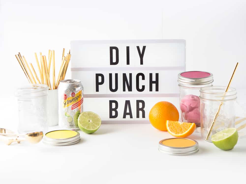 A white lightbox sign with words, "DIY Punch" Bar surrounded by cans of Schweppes sparkling water, mason jars, gold straws, and slices of fruit.