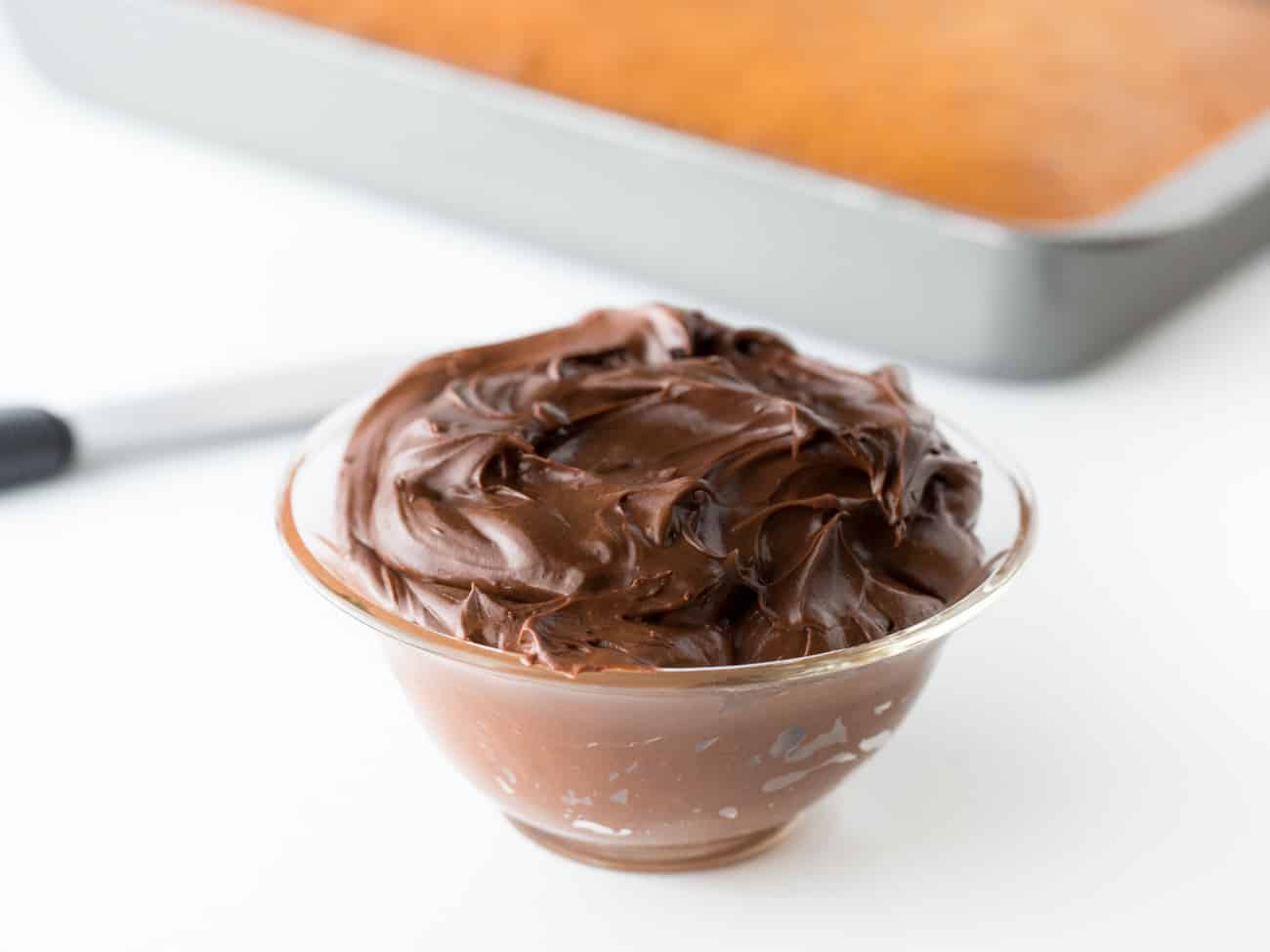 Plastic bowl of easy chocolate frosting with chocolate spreader and 9x13 yellow cake in background.
