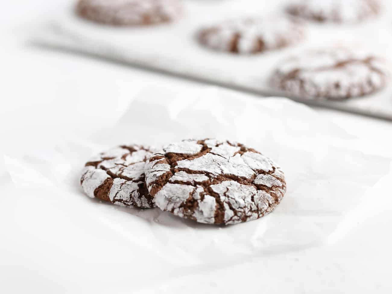 Two crackle top chocolate brownie cookies with baking tray in background.